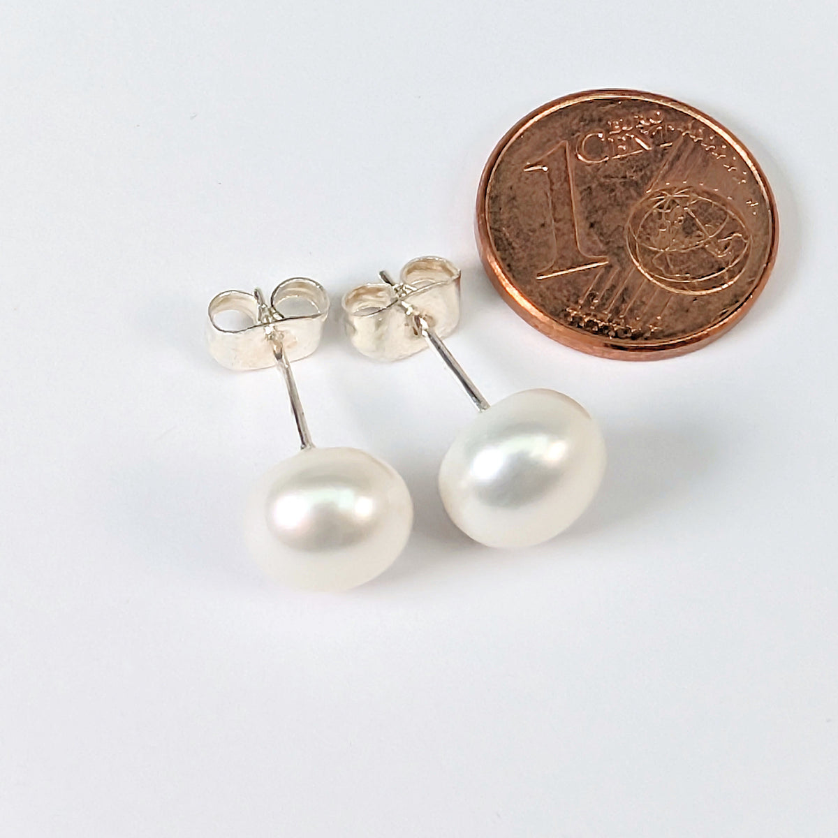 Paire de boucles d'oreilles Diana en perle de culture blanche de 8mm, monture clou en argent 925, posées à côté d'une pièce de 1 centime pour comparer la taille