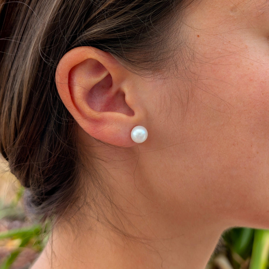 Femme portant une boucle d'oreille Diana à l'oreille, clou en plaqué or 18K surmonté d'une perle de culture blanche en forme de bouton d'environ 8mm, vue de profil sur fond naturel