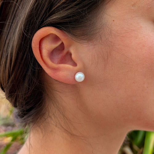 Femme portant une boucle d'oreille Diana à l'oreille, clou en plaqué or 18K surmonté d'une perle de culture blanche en forme de bouton d'environ 8mm, vue de profil sur fond naturel